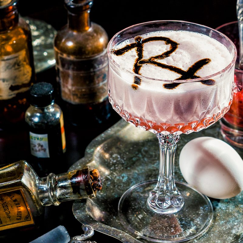 A close-up of a cocktail with "Rx" beside an egg, a glass filled with red substance, and bottles of alcoholic beverages.