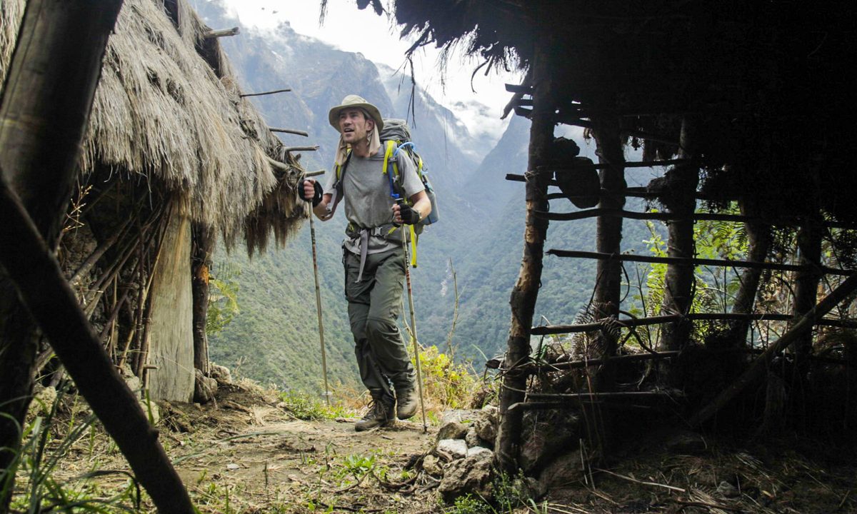 A man with backpack holding two trekking polls approaching a hut in the mountains.