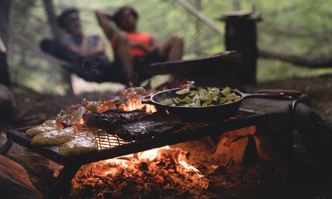 Camping food grilling over an open flame with a blurred couple seated in the background