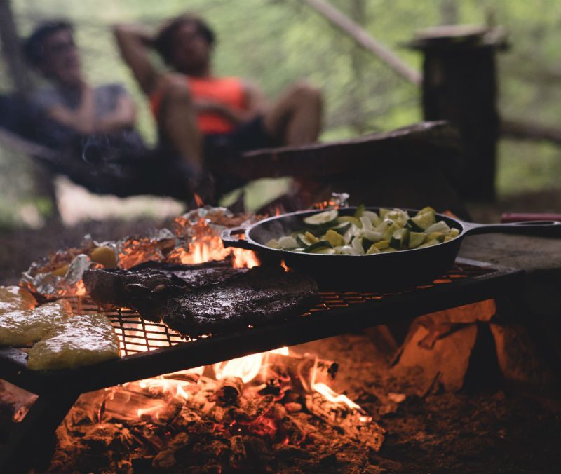 Camping food grilling over an open flame with a blurred couple seated in the background