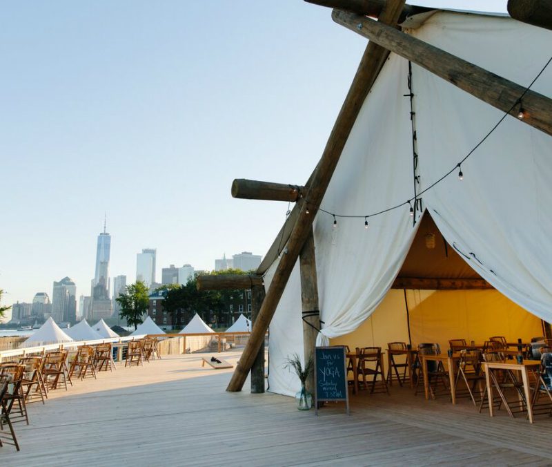 A large event tent is set up on an expansive deck on Governor's Island overlooking New York harbor with the skyline in the distance. 