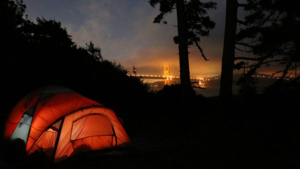 A tent glows from within in a night shot with the Golden Gate Bridge in the distance. 