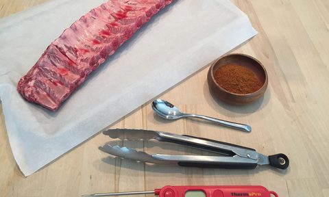 Raw ribs with tongs, a dry rub, a meat thermometer, and a spoon