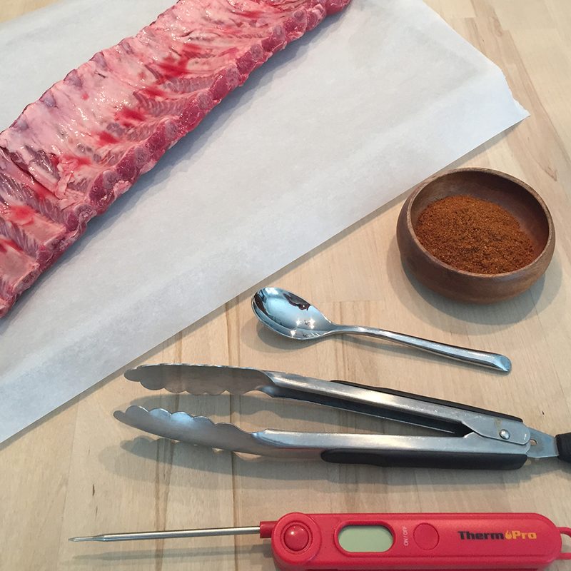 Raw ribs with tongs, a dry rub, a meat thermometer, and a spoon