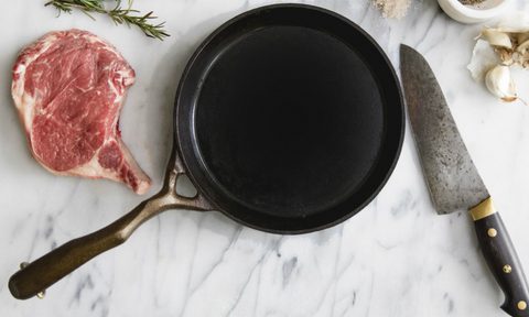 Cast-iron skillet on marble slab with steak and knife.