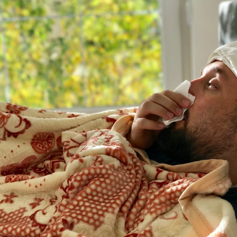 Man sick in bed, blowing his nose into a tissue.