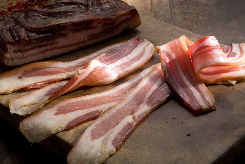 A slab of bacon and strips of raw bacon on a wooden surface.