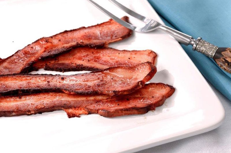 A plate of bacon, two-tine fork, and a blue napkin on a smooth surface.