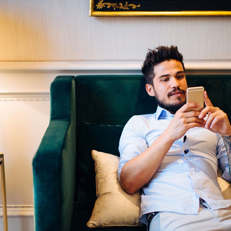 A man slouches on a couch while looking at his cell phone.