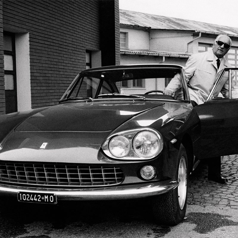 Enzo Ferrari getting out of a Ferrari in front of brick buildings.