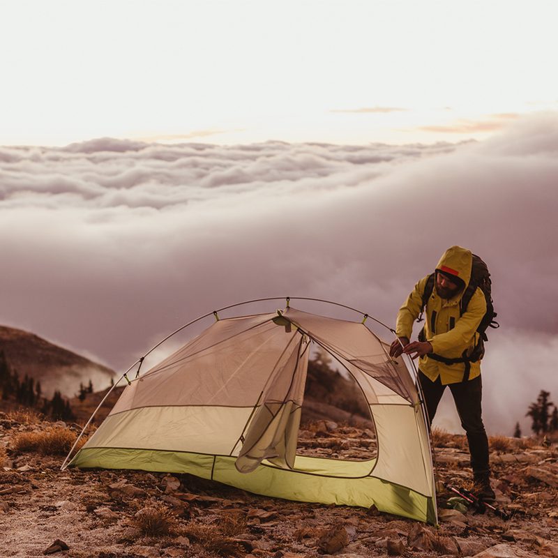 A man wearing a hooded jacket attempting to set up a tent in the wilderness.
