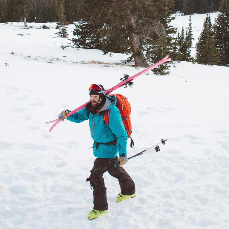 Man trekking through snow carrying skis and ski poles.