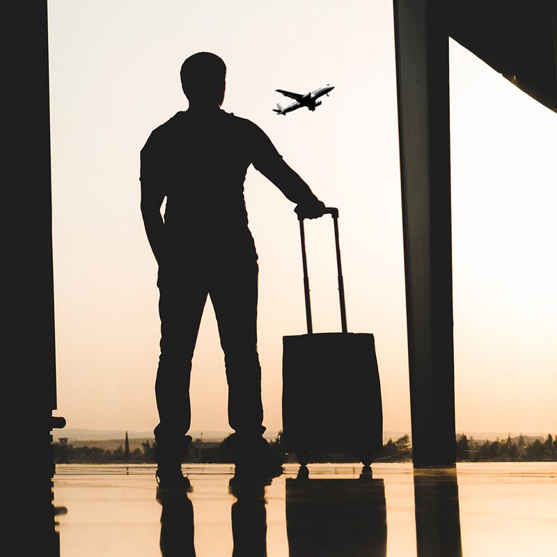 Man with a suitcase watching an airplane take off through the airport window.