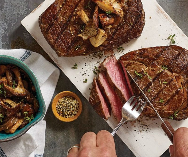 pair of hands slicing steak with mushrooms and herbs on top.