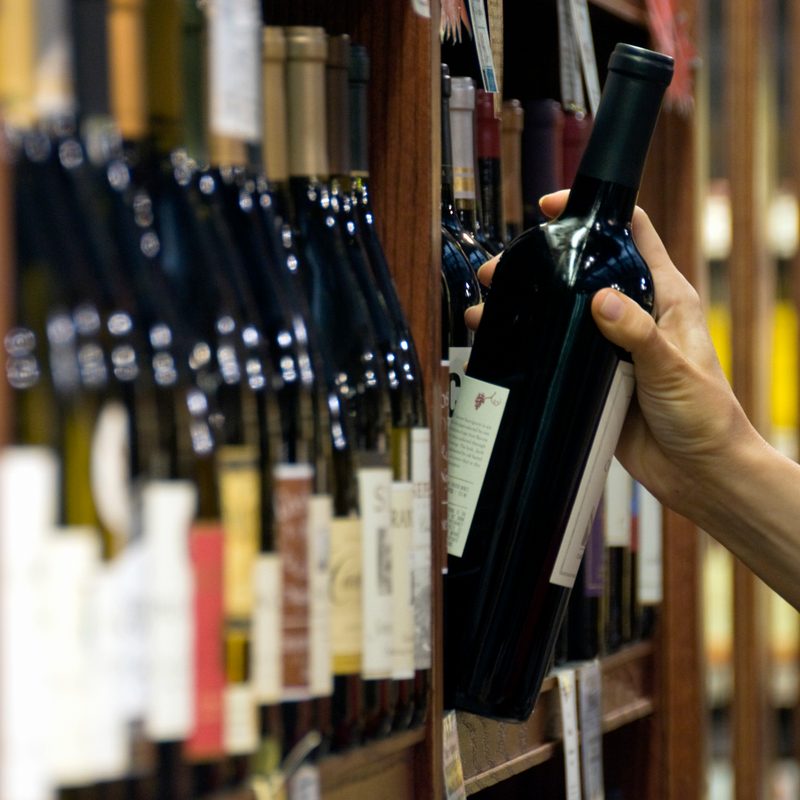 A woman's hand reaches out to select a bottle of red wine from the shelf of a wine shop
