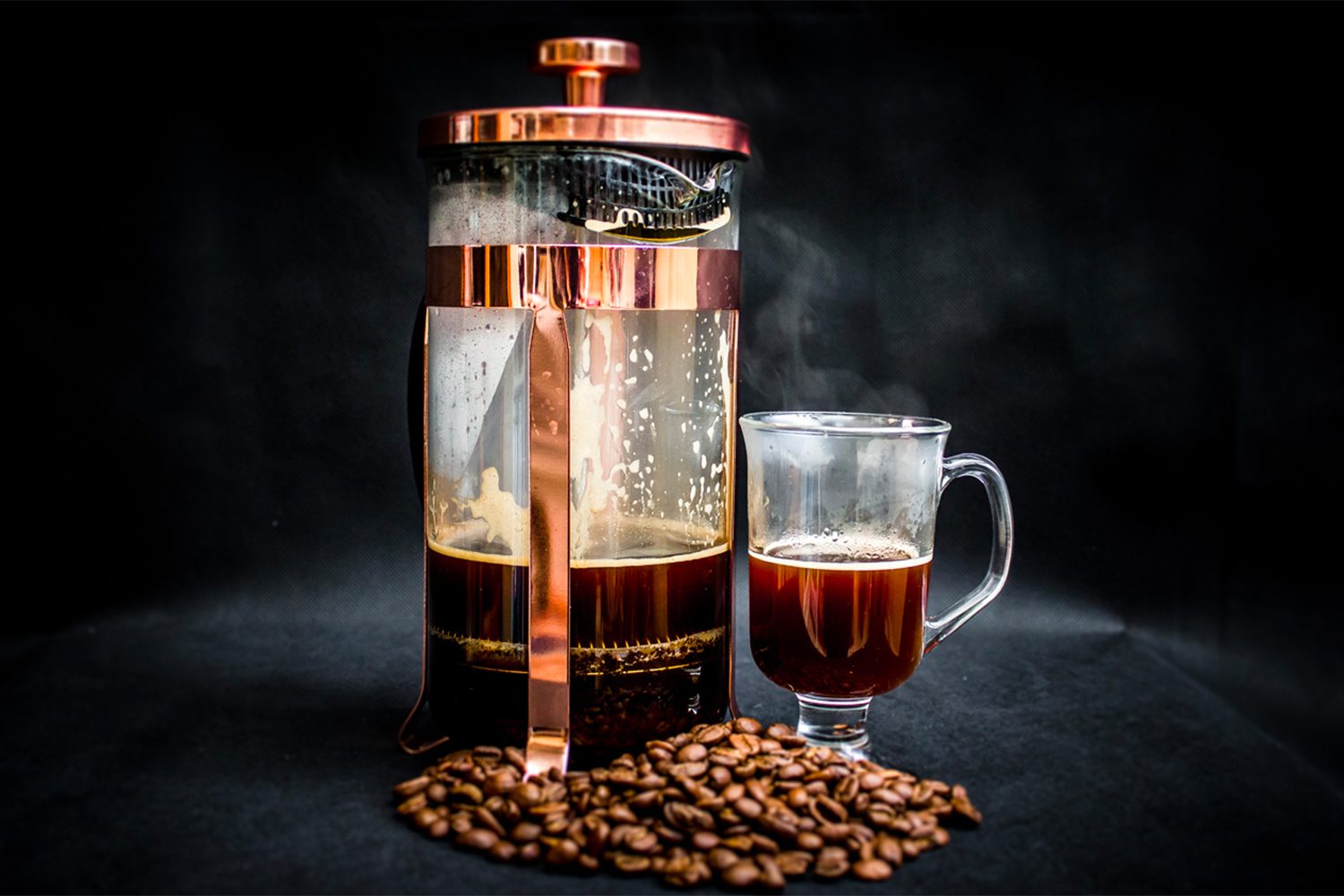 A French press coffee maker next to a glass of coffee, sitting beside a pile of coffee beans on a black table