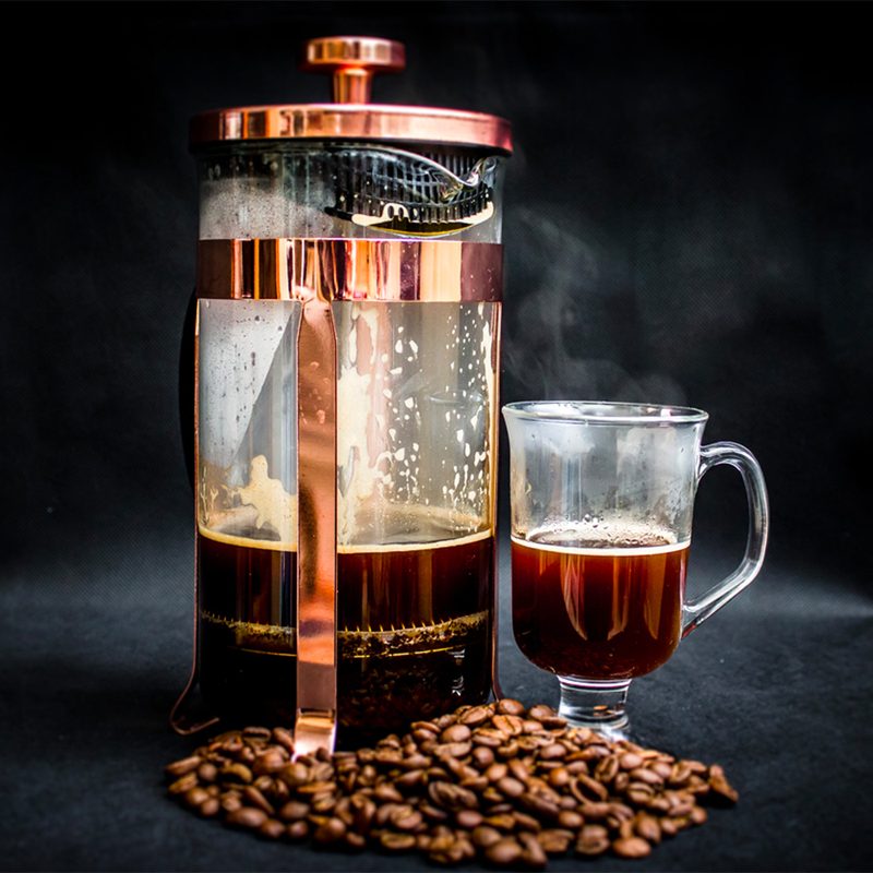 A French press coffee maker next to a glass of coffee, sitting beside a pile of coffee beans on a black table