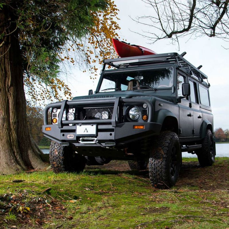 Classic Land Rover Defender parked in between two trees with a lake in the background.
