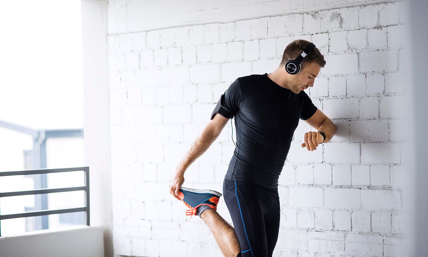 Man wearing headphones as he leans on a wall and stretches a single leg.