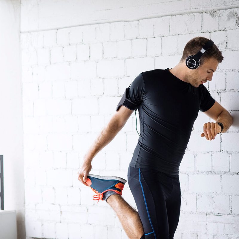 Man wearing headphones as he leans on a wall and stretches a single leg.