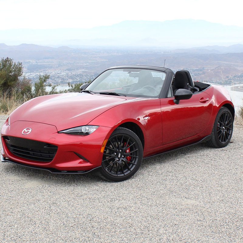 Red Mazda MX-5 Miata parked on the side of a gravel road in front of a hill.