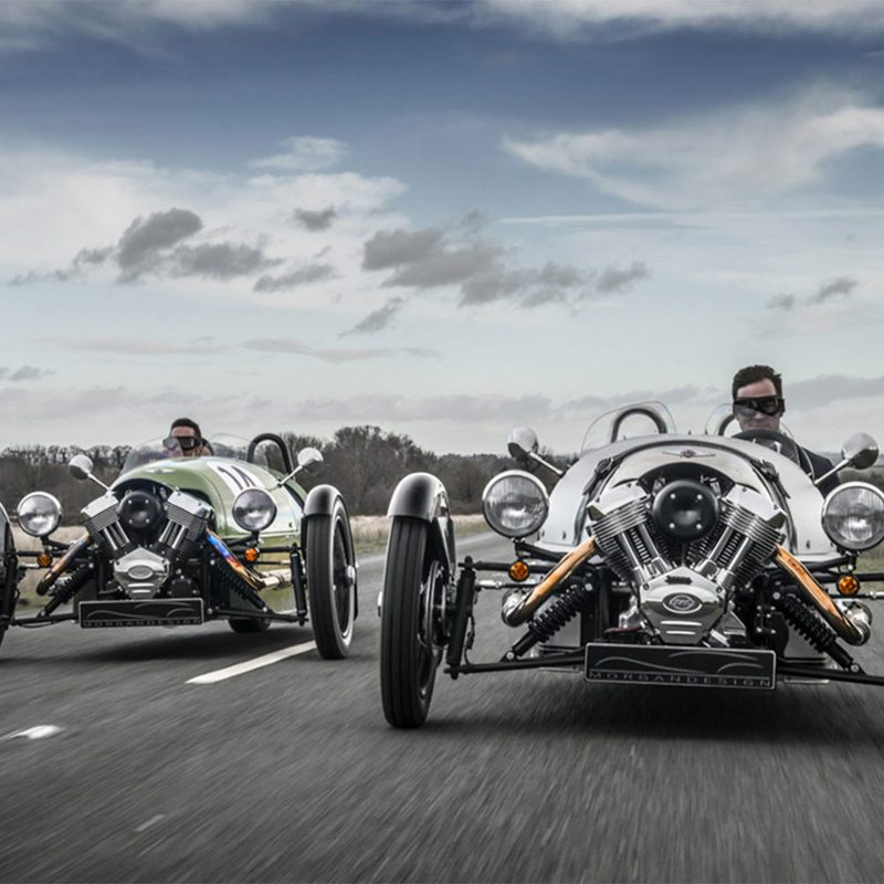 Two Morgan 3-Wheelers driving on a road during a cloudy day.