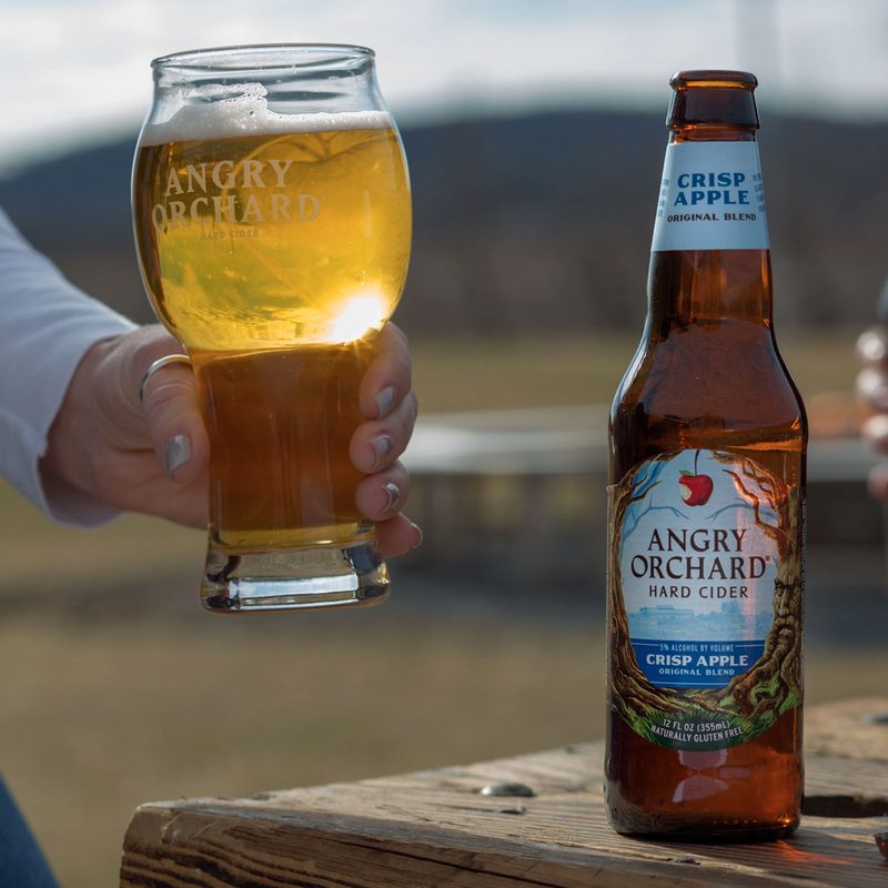 An Angry Orchard Hard Cider bottle in Crisp Apple along with a person holding a glass filled with cider.
