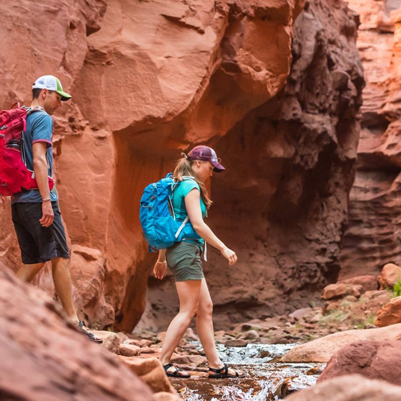 Two hikers in summer clothing cross a small creek in the red bedrock