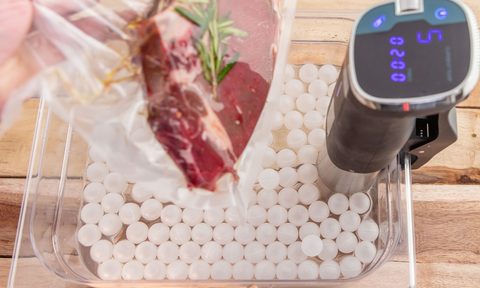 A hand holding a vacuum-sealed bag containing meat on a tray with a sous vide machine