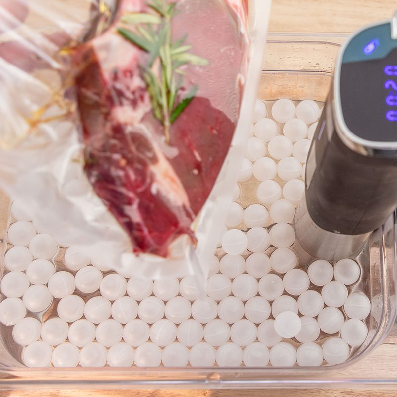 A hand holding a vacuum-sealed bag containing meat on a tray with a sous vide machine.