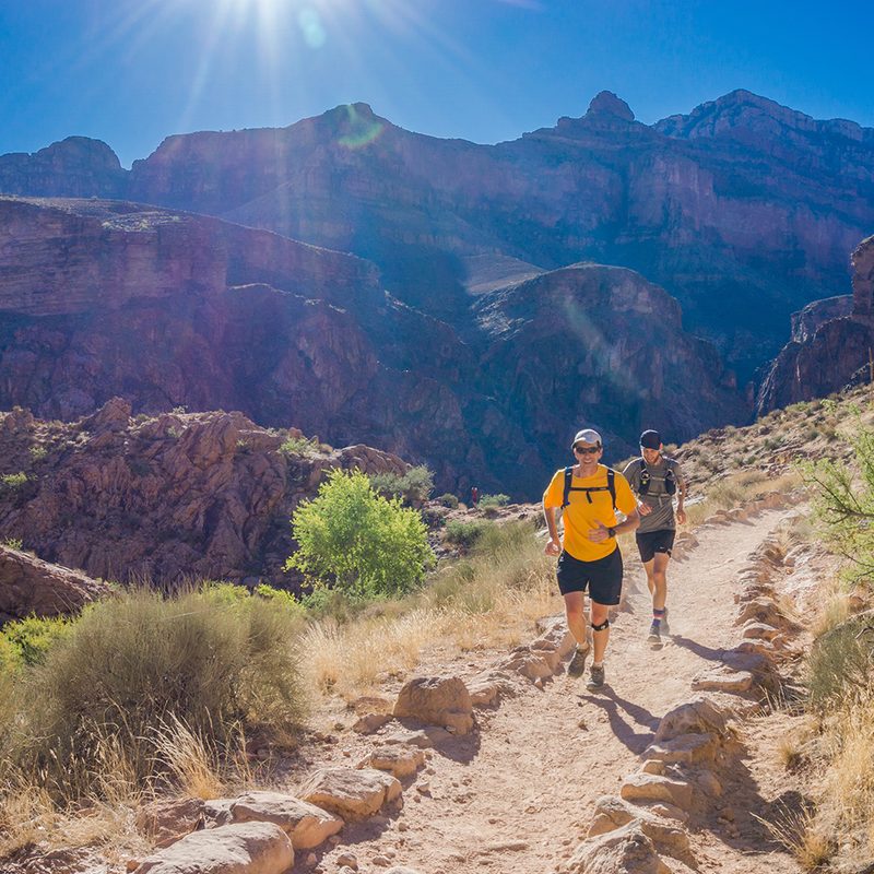 Two runners run on a trail with mountains in the background.