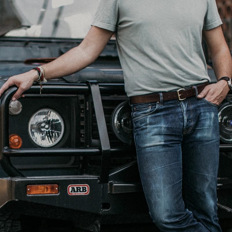 Torso shot of a man wearing jeans and a leather belt while holding onto his vehicle.