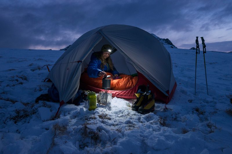 Woman with a headlamp camping in a winter tent surrounded by snow.