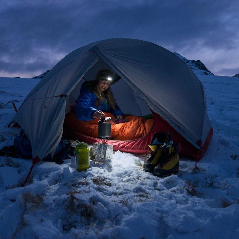 Woman with a headlamp camping in a winter tent surrounded by snow.