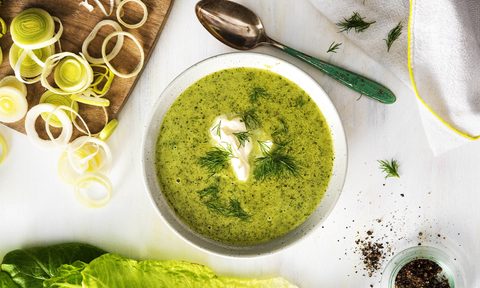A bowl of lettuce leek soup next to a spoon and cutting board with sliced leeks.