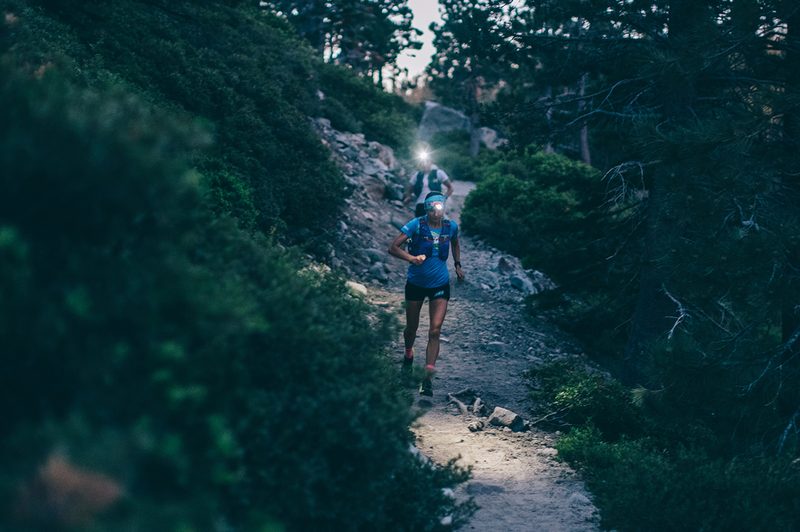 Two runners run downhill on a track between forests, their headlamps illuminate the trail.