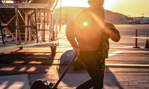 Traveler pulling a rollaboard suitcase outside an airport, sun shining directly into the camera.