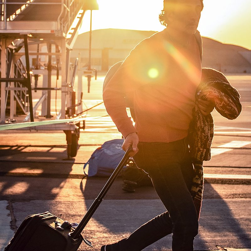 Traveler pulling a rollaboard suitcase outside an airport, sun shining directly into the camera.