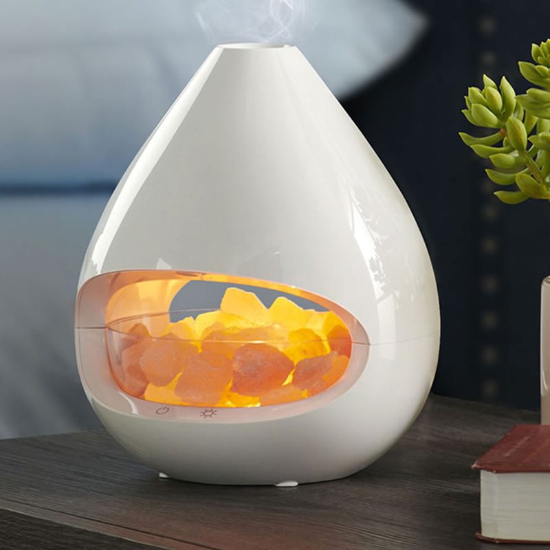 A Hammacher Schlemmer Himalayan Salt Lamp Diffuser beside a plant box and eyeglasses on top of a thick book on a wooden bedside table.