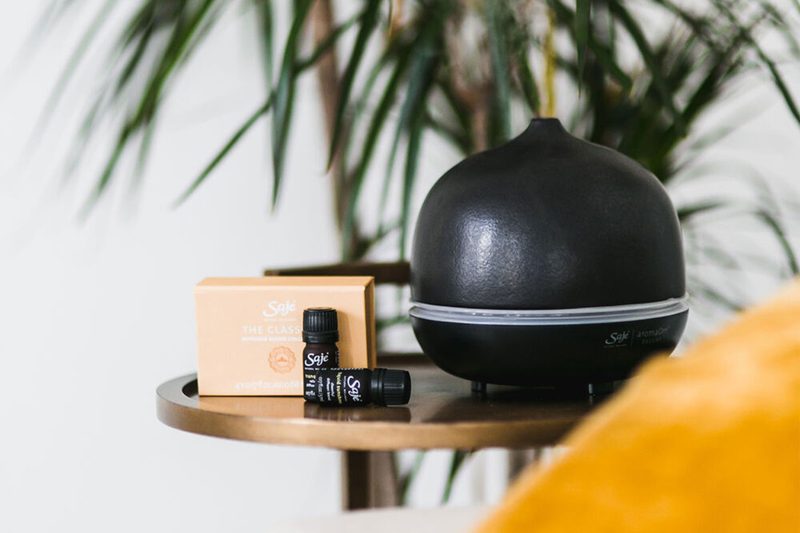 The Saje Aroma Om Deluxe Diffuser beside two bottles of essential oils and a box on a wooden round table.