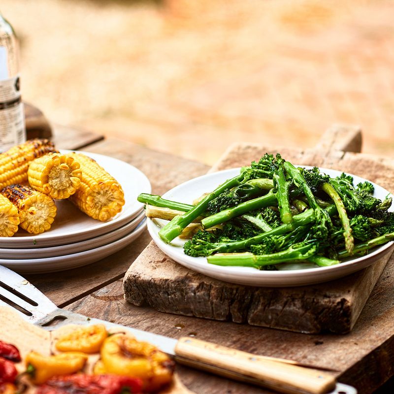 A plate of grilled corn, a plate of grilled broccolini, and grilled peppers on the side.