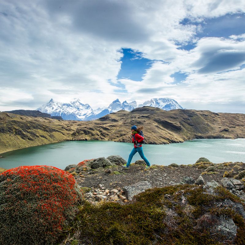 Hiker in a red coat walking with a backpack near a backcountry lake, mountains in the background.