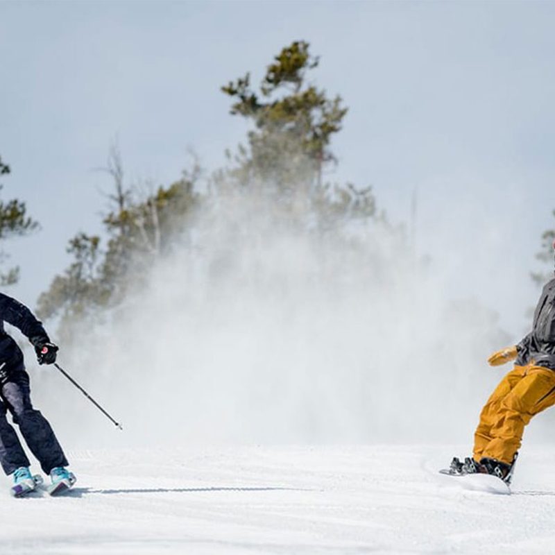 A skier and a snowboarder coming down a snowy mountain together toward the camera.