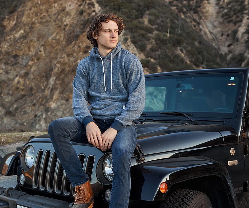 A man sitting on his car with a hoodie, staring off into the distance.