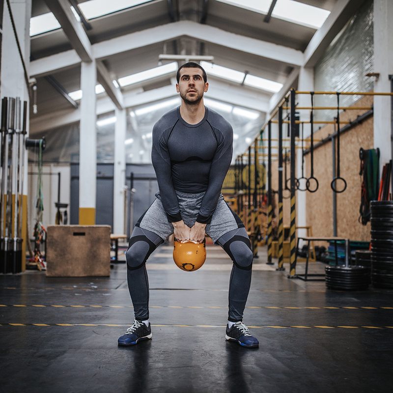 A man who's holding a kettlebell as he bends his hips and knees to perform kettlebell swings at the gym.