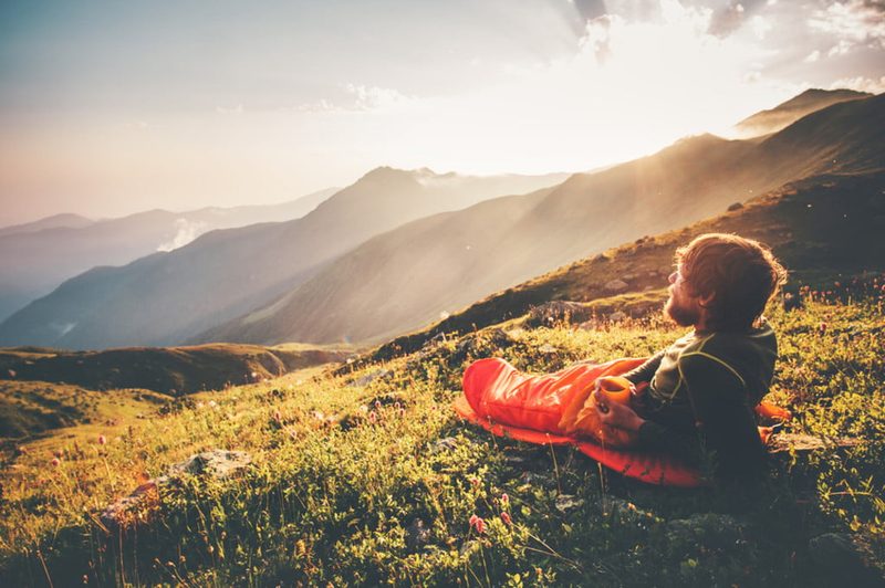 A man lays in a sleeping bag on the grass, looking out over the mountains as the sun sets