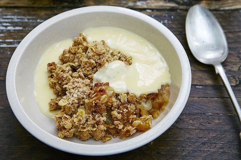 A bowl of classic apple crumble beside a spoon on a wooden table.