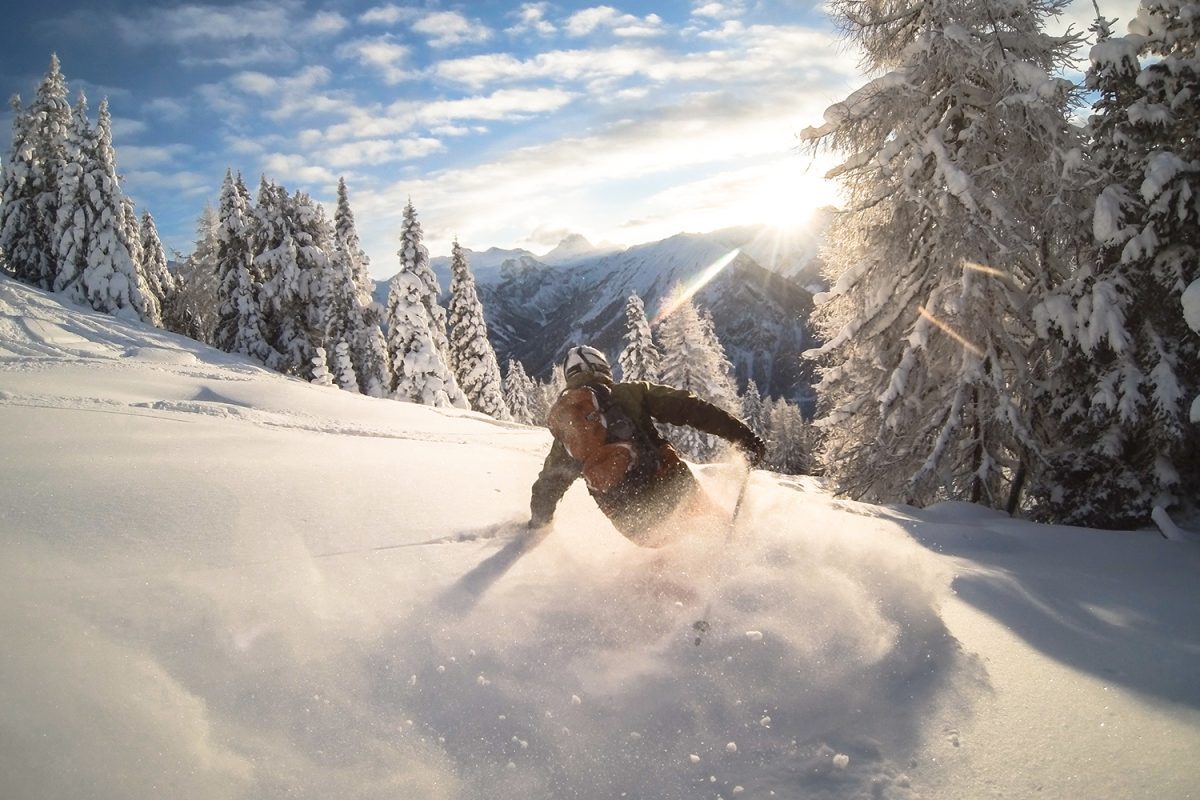 A man puts in a deep powder turn near some pine trees that are covered In snow. In the background, the sun sets over some mountains.