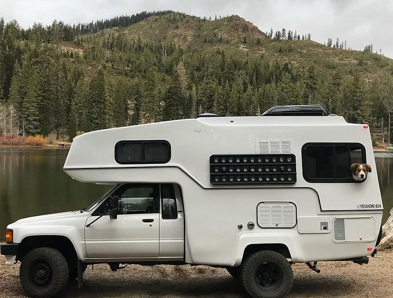 Modern white truck camper near a lake with a dog peering through the rear window.