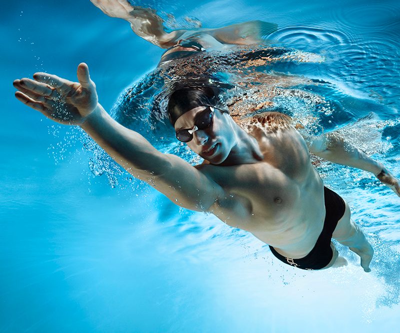 Man swimming while wearing a swim cap, goggles, and shorts.